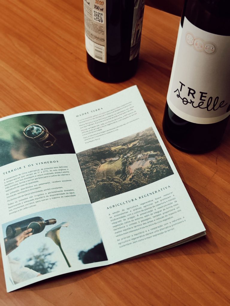 An open informational booklet written in Portuguese sits on a wooden tabletop next to two dark wine bottles, one featuring a label that reads "Tre Sorelle."