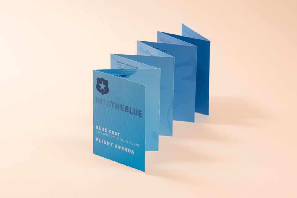 A blue accordion-folded event guide standing expanded in a zigzag shape on a light surface, titled "INTO THE BLUE - Blue Coat 2009 Worldwide Sales Summit Flight Agenda."