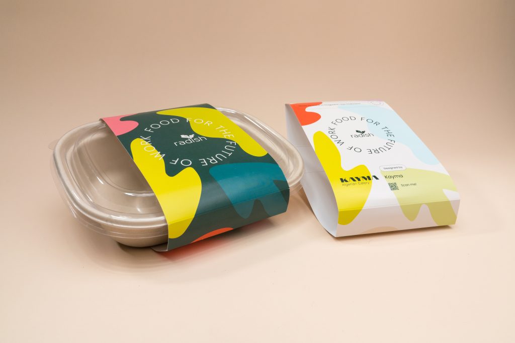 An off-white plant-fiber food container with a clear plastic lid wrapped in a colorful packaging sleeve, sitting beside a standalone folded paper sleeve against a plain, light beige background.