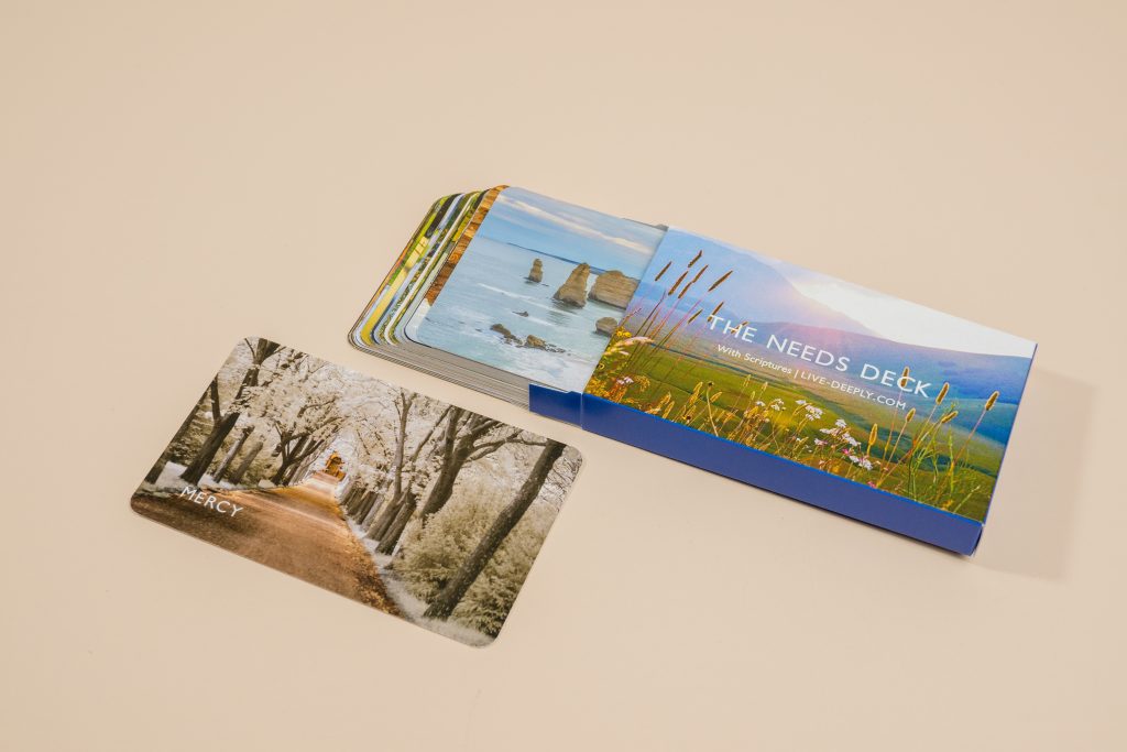 A deck of prompt cards sliding out of a blue custom sleeve, with a single card pulled out in front showing a tree-lined path labeled "Mercy."