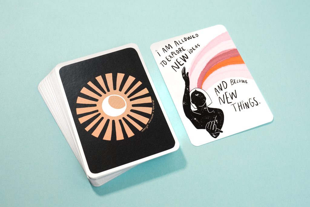 A stacked deck of black cards with a sun-burst design placed next to an affirmation card reading "I am allowed to explore new ideas and become new things" on a light teal background.