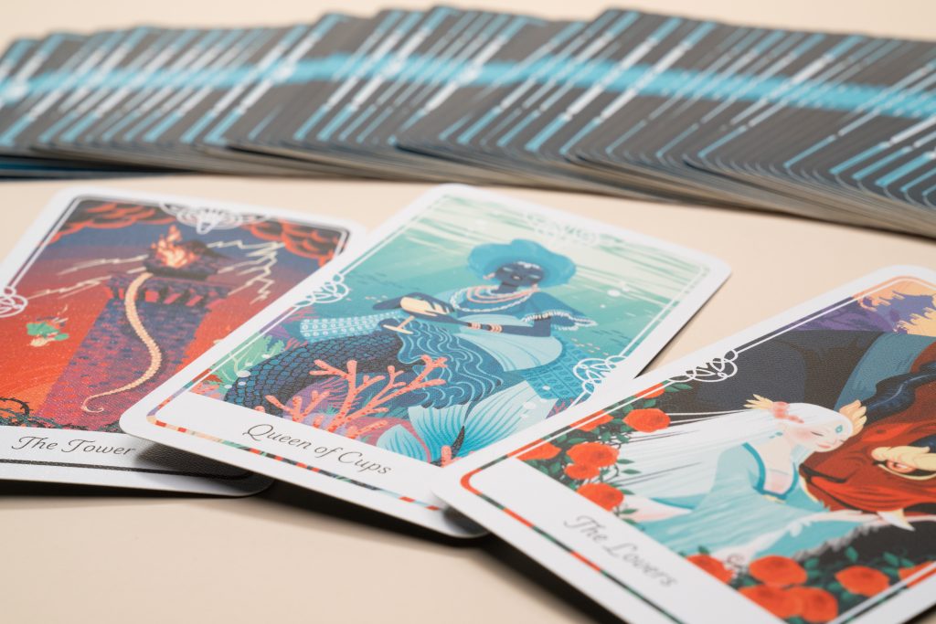 A close-up view of illustrated tarot cards spread out on a beige surface, featuring cards labeled "The Tower," "Queen of Cups," and "The Lovers" in the foreground with a fanned deck of card backs behind them.