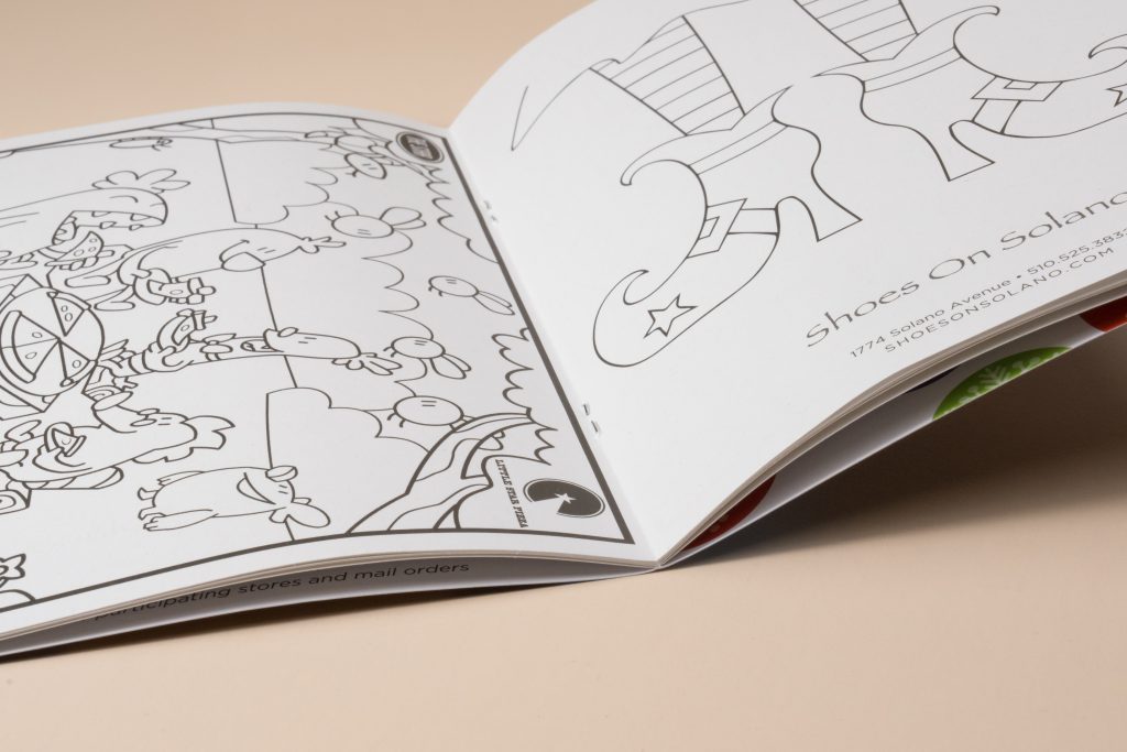 An open black-and-white activity book resting on a light beige surface, featuring line art drawings and the text "Shoes On Solano" on the right page.