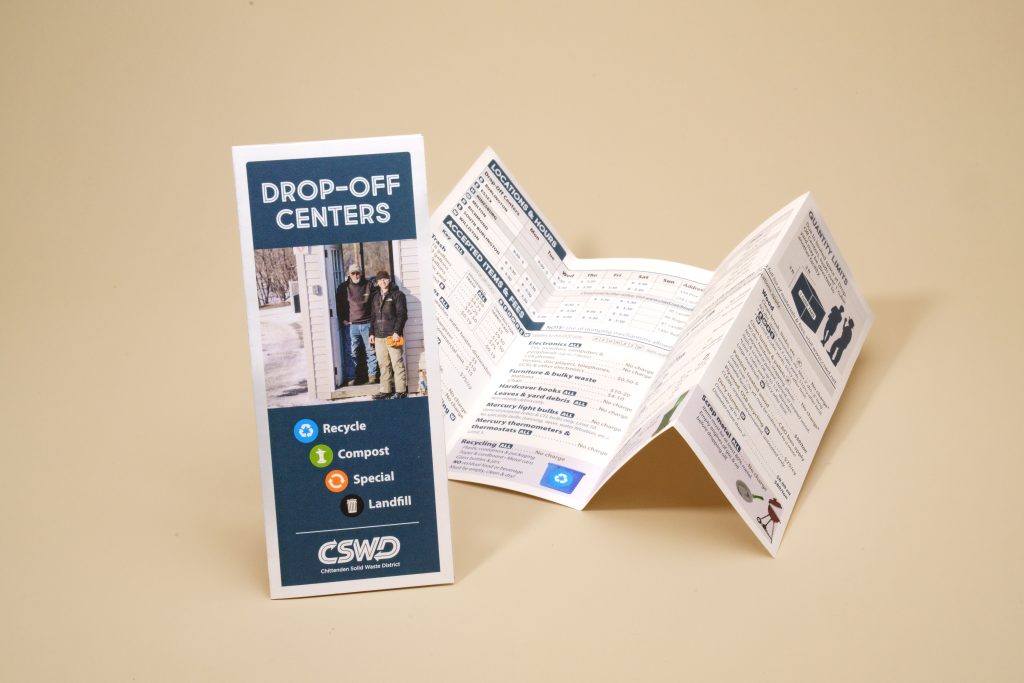 A folded informational pamphlet titled "Drop-Off Centers" standing next to an accordion-folded brochure outlining waste management schedules and accepted items on a tan surface.